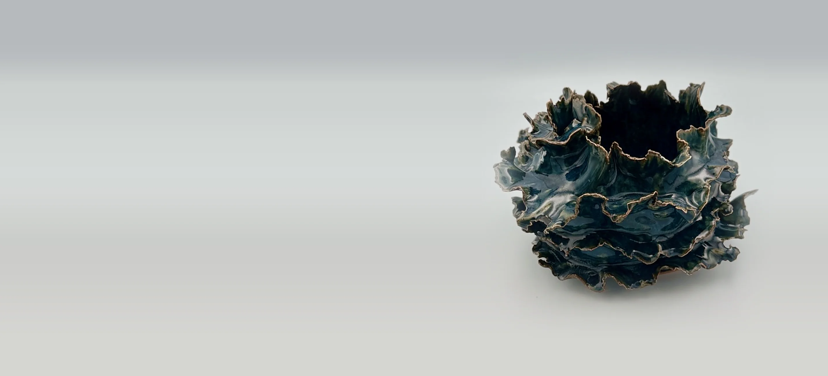 A Krysta Mae ruffled sculpture in deep blue-green glaze with bronze edges, on a soft grey surface
